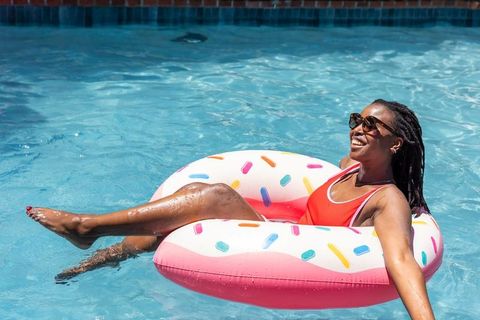 Joyful african woman lounging on pink inflatable donut ring in sunny pool wearing red swimsuit