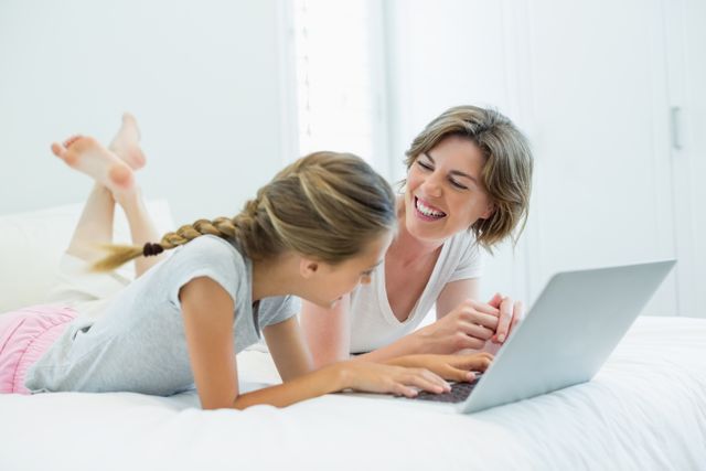 Mother and daughter enjoying quality time together while using a laptop on a bed in a bright bedroom. Perfect for illustrating family bonding, technology use in daily life, and parent-child relationships. Ideal for use in parenting blogs, family lifestyle articles, and technology-related content.