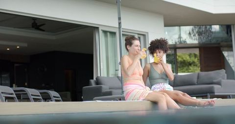 Two friends relaxing on modern poolside patio sipping cold drinks in swimwear and towels