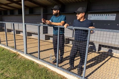 Two men standing in dugout leaning on wire fence watching baseball game during sunny day