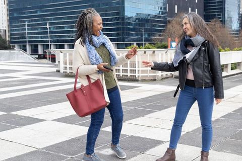 Laughing senior women exchanging keys on urban plaza with red tote and city skyline