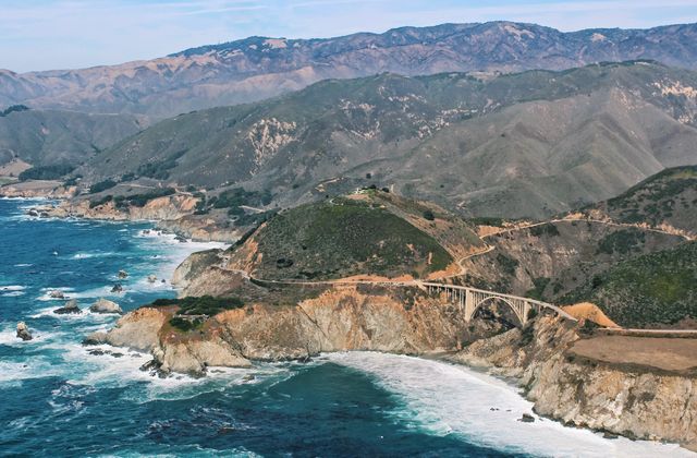 Aerial view of Big Sur coastline showcasing the iconic Bixby Creek Bridge against a backdrop of rugged mountains and the Pacific Ocean. Useful for travel blogs, tourism promotions, digital postcards, and nature magazines emphasizing scenic views and natural beauty spots in California.
