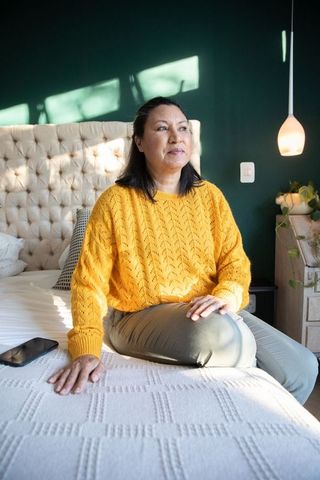 Mature woman sitting on sunlit bedroom bed wearing mustard sweater and olive trousers