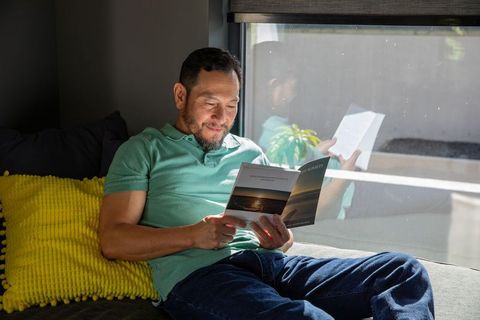 Asian man reading paperback on sofa by window with yellow cushion in sunny interior