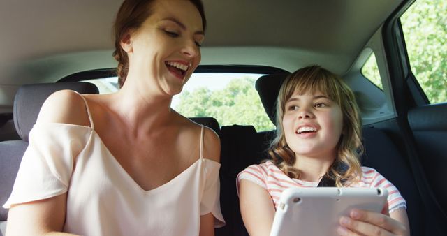 Mother and daughter enjoying time together in the back seat of a car, using a tablet. They are both smiling and appear to be having a good time. This image can be used for family travel, technology use in family settings, parental bonding, and happy moments. Ideal for articles or advertisements focusing on family dynamics, digital education, or family vacations.