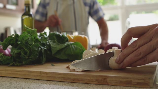 A senior woman actively chops vegetables on a wooden board, while her husband stirs a pot in the background, symbolizing teamwork and togetherness. Ideal for portrayals of simplicity, healthful lifestyles, or at-home activities for elderly during quarantine periods. Suitable for articles on healthy recipes, lifestyle pieces on senior couples, or promotions for kitchenware products engaging older demographics.