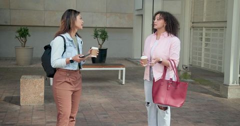 Two african women chatting over takeaway coffee in urban courtyard with red tote