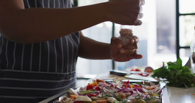 Close-up depicts person seasoning colorful mixed vegetables on baking tray in a modern kitchen. Great for promoting healthy eating, diverse representation, and inclusivity in culinary arts. Useful in blogs, digital ads, and marketing materials focused on cooking, lifestyle, and wellness.