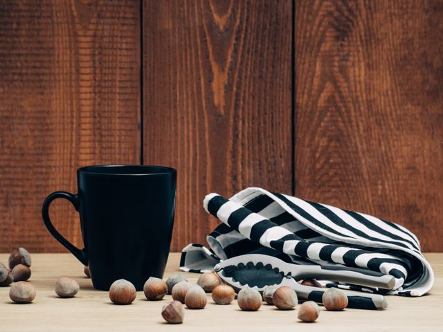 Black coffee mug placed on rustic wooden table with scattered hazelnuts, accompanied by black and white striped kitchen towel and nutcracker. Perfect for concepts related to rustic kitchen decor, coffee drinking moments, warmth and coziness, culinary themes, and country-style kitchen settings.