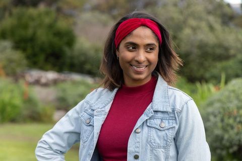 Indian woman in 20s smiling and looking left in garden wearing denim jacket and red headband