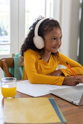 Preteen girl studying with headphones and laptop, writing in notebook beside orange juice