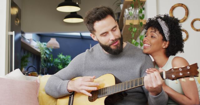 Image of happy diverse couple relaxing in living room, man playing guitar and signing to woman. Happiness, inclusivity, free time, romance, music, togetherness and domestic life.