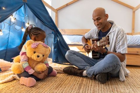 Indian father playing guitar while african american daughter hugs teddy in cozy tent
