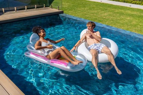 Young couple relaxing on inflatable floats in sunny backyard pool holding drinks and smiling