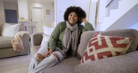 Smiling woman relaxing on gray sofa in cozy living room touching hair wearing casual green sweater