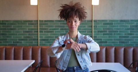 Young black woman standing in cafe wearing denim jacket and smartwatch glancing at camera