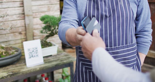 Customer making a contactless digital payment to a vendor at a local farmers market. Vendor wears apron and holds device while other hand has smartphone for transaction. Background includes plants and table with QR code. Use for themes related to digital transactions, small business operations, sustainable shopping, and cashless payments.