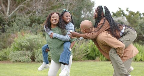 African American family giving piggyback rides and laughing on grassy park lawn