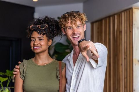 Smiling mixed couple posing in modern lobby, man pointing, woman holding bag strap
