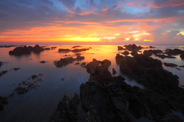 Stunning seaside coastline during sunset. Features rocky formations extending into calm waters with a vibrant, dramatic sky in the background. Ideal for travel blogs, nature magazines, relaxation themes, and inspirational content promoting tranquility and the beauty of nature.