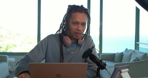 Mid-adult african american man livestreaming with laptop and mic overlooking ocean view