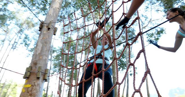 A young Caucasian girl is climbing a rope net in an outdoor adventure park, with copy space. She is being assisted by a woman, engaging in a team-building exercise or recreational activity.
