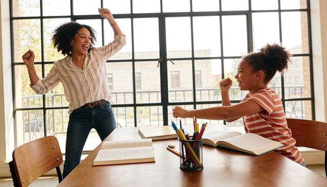 Mother and daughter celebrating homework success at sunlit table by large grid window