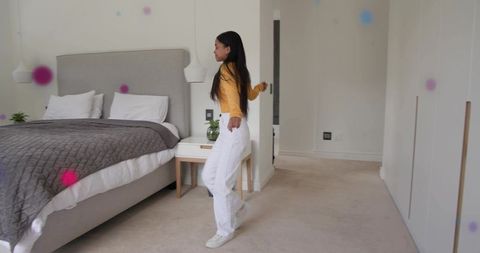 Asian woman standing in minimalist bedroom wearing mustard top and white pants near wardrobe