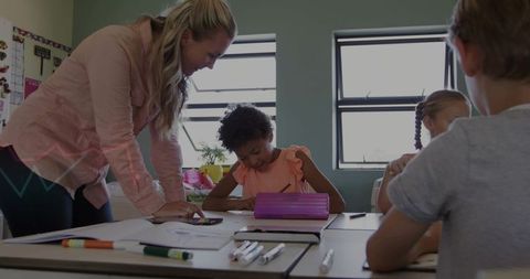 Teacher Leaning Over Student Guiding Writing at Classroom Table with Tablet and Pencil Case