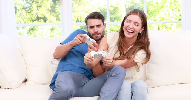 Young couple sitting on a sofa in a living room, playing video games using controllers. They appear to be having fun and enjoying themselves. Ideal for advertisements, blog posts on relationships, gaming, leisure activities, or home decor websites.