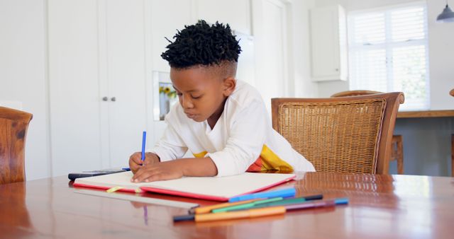 Young boy is concentrating while drawing with crayons in a brightly lit kitchen, promoting the themes of creativity and education. Ideal for use in educational content, parenting blogs, creative projects, or home lifestyle articles.