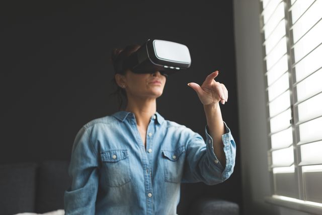 Woman wearing a virtual reality headset, interacting with digital content in a modern home. Ideal for illustrating concepts of technology, innovation, and immersive experiences. Suitable for use in articles, blogs, and advertisements related to VR technology, gaming, and modern lifestyles.