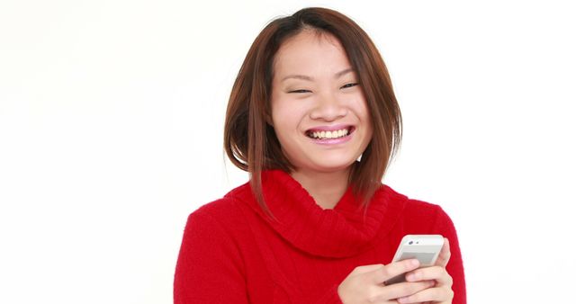 Asian woman smiling while using smartphone in red sweater captures moment of joy and technology. Perfect for illustrating communication, technology use, happiness, and lifestyle in advertisements or blog posts.