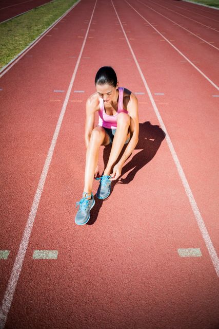 Female athlete tying her running shoes on a running track, preparing for a workout or race. Ideal for use in sports and fitness advertisements, training programs, health and wellness articles, and motivational content.