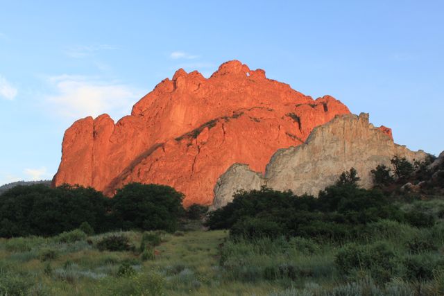 A striking red rock formation illuminates in the golden hour, creating a vivid contrast with the lush green vegetation below and the clear blue sky above. Ideal for use in travel brochures, nature-themed calendars, postcards, or websites showcasing natural landscapes. Emphasizes natural beauty, outdoor adventure, and exploration.