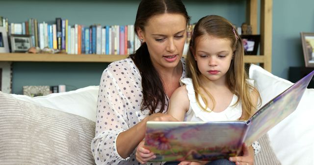 Mother reading book to daughter while sitting on a comfortable sofa, close to a bookshelf. Ideal for themes related to family bonding, education, parenting, and cozy home environments. Can be used in advertisements, blogs, family magazines, and educational content featuring parent-child learning activities.