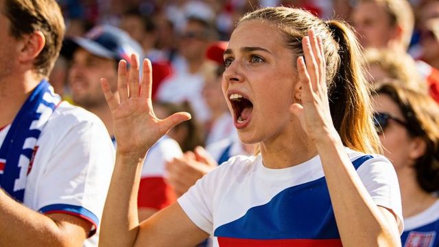 Young female fan shouting and cheering in packed stadium wearing blue and red jersey