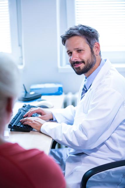 Doctor engaging with patient while using computer in medical office. Ideal for healthcare, medical consultation, patient care, and hospital-related content.