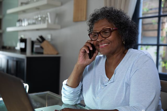 Senior African American woman sitting in her kitchen, using a laptop and talking on the phone. She is smiling and appears to be engaged in a pleasant conversation. This image can be used to depict themes of elderly people embracing modern technology, remote communication, and domestic life. Ideal for articles or advertisements related to senior citizens, technology use among the elderly, or home-based activities.