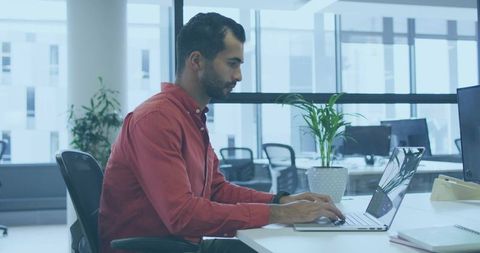 Middle eastern man working on laptop in modern office workspace, casual red shirt