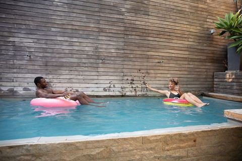 Diverse couple floating on colorful inflatable tubes in sunny backyard pool with cocktails