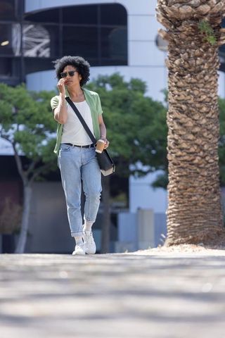 Walking through city plaza adjusting sunglasses while holding coffee and crossbody bag