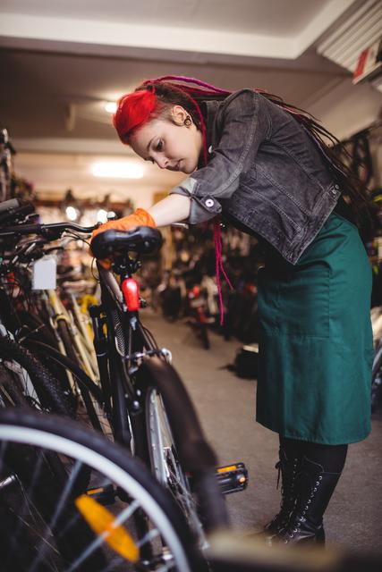 Image shows a female mechanic with red hair and dreadlocks examining a bicycle in a workshop. She appears focused and dedicated to her work. This image can be used for content related to bike repair services, professional mechanics, women in skilled trades, and bicycle maintenance tutorials. It conveys themes of dedication, precision, and technical expertise.