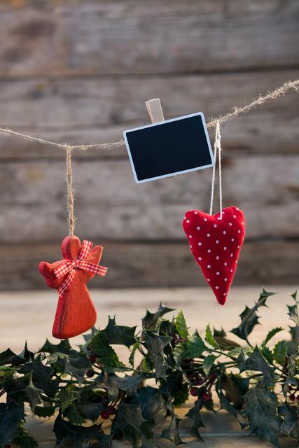 Christmas decorations including a red angel and a polka dot heart hanging on a string against a rustic wooden background. Holly garland is visible at the bottom. Ideal for holiday greeting cards, festive invitations, seasonal blog posts, and Christmas-themed advertisements.