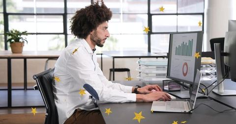 Middle Eastern man typing at office desk analyzing dashboard charts on dual screens