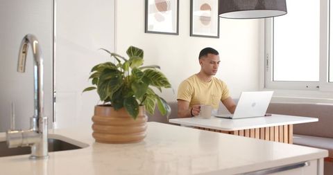 Man working from home at minimalist kitchen nook holding coffee mug with laptop