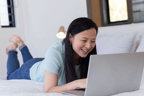 Asian woman smiling while using laptop on bed in minimalist bedroom for remote work