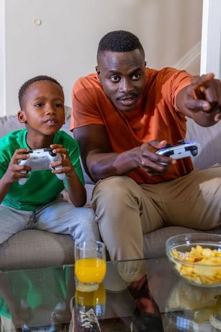 African american father and son playing video games on couch, bonding through friendly console play