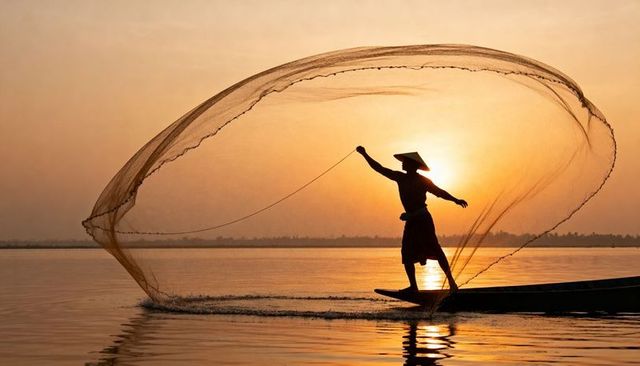 Silhouette fisher casting circular net from boat at golden sunset with conical hat