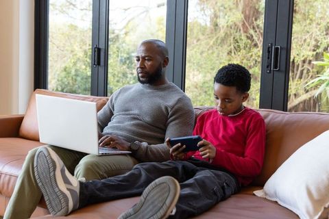 African american father working on laptop while son playing on handheld device on sofa